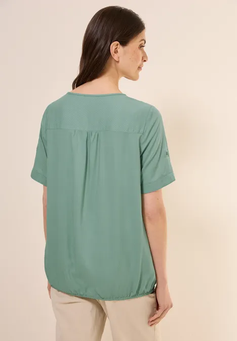 Frau mit grünem Kurzarmshirt von hinten, trägt beigefarbene Hose.